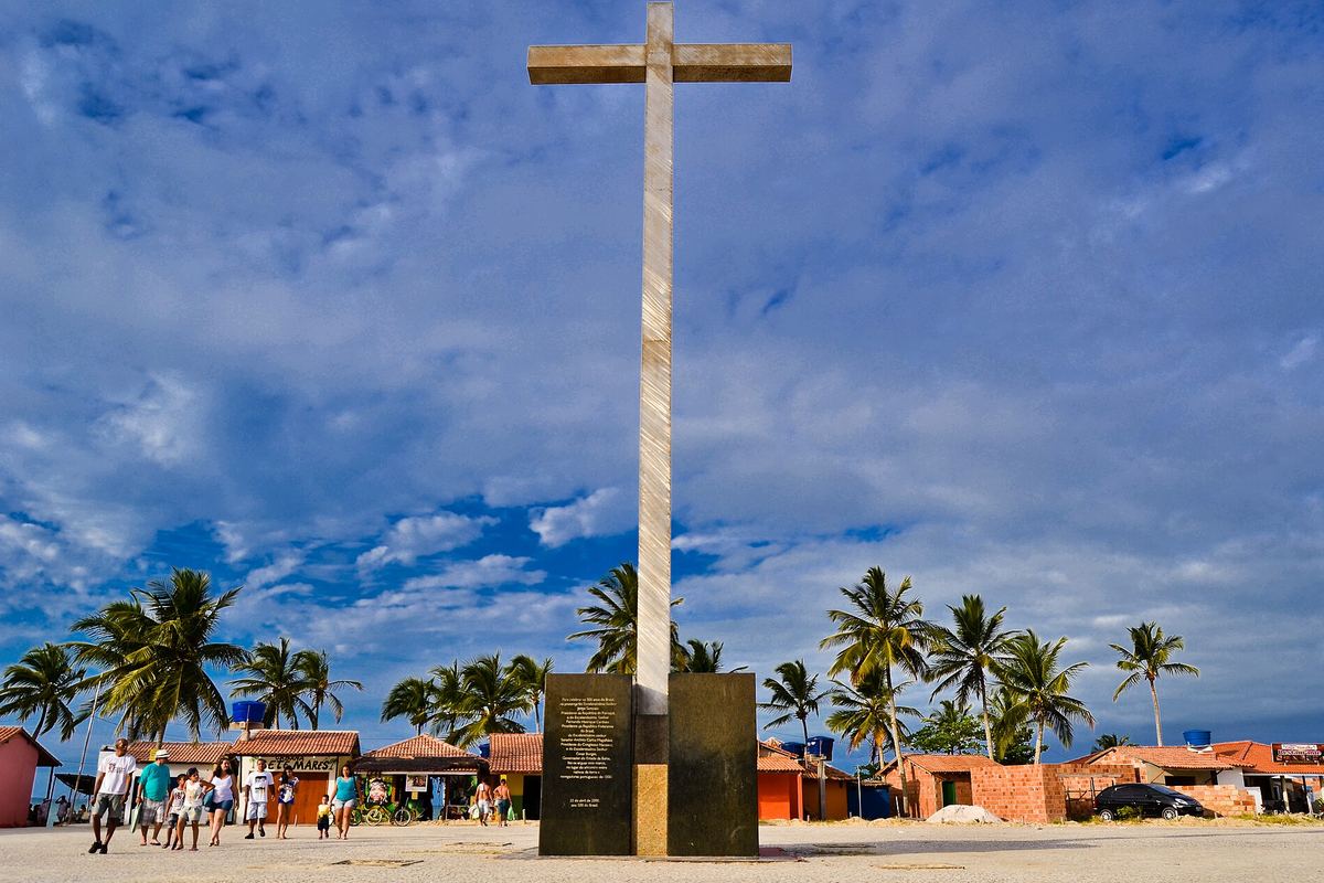 Cruz de pedra erguida sobre um pedestal na praia de Coroa Vermelha, com coqueiros e casas ao fundo